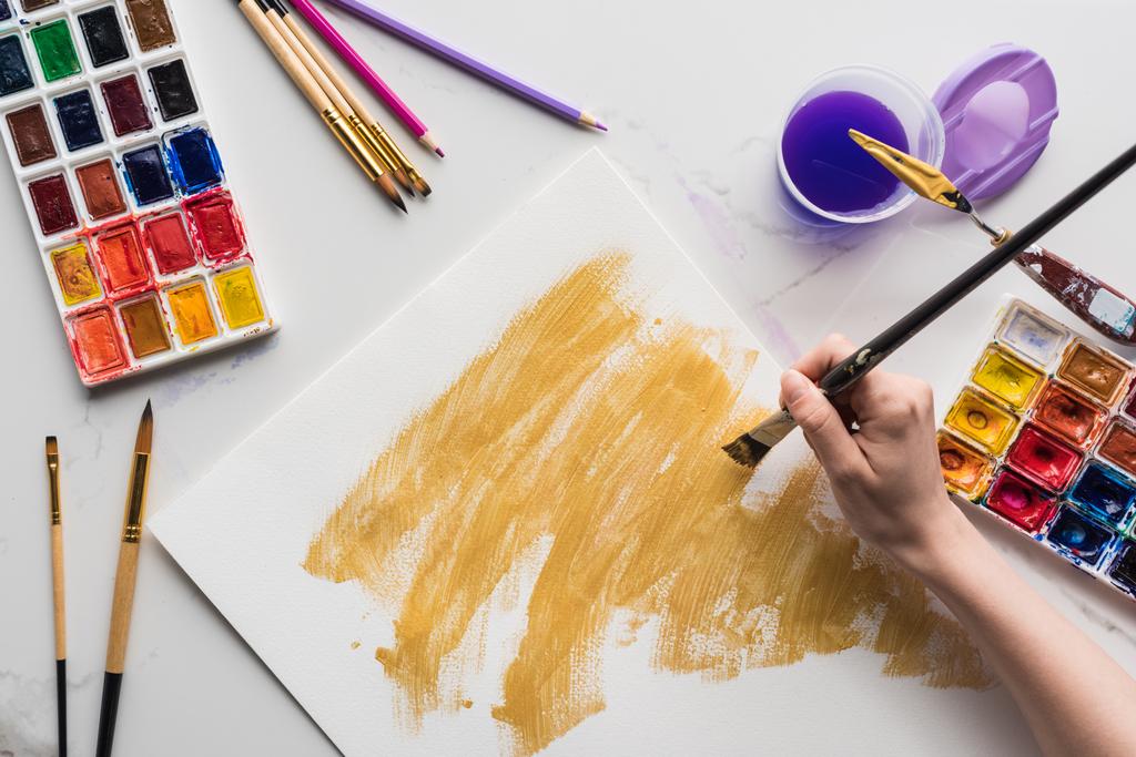 bijgesneden beeld van kunstenaar tekening gele aquarel penseelstreken op wit papier op marmer wit oppervlak - Foto, afbeelding
