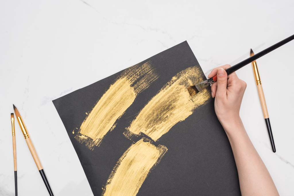 Teilansicht des Künstlers beim Zeichnen von Pinselstrichen mit goldener Farbe in schwarzem Papier auf Marmoroberfläche - Foto, Bild