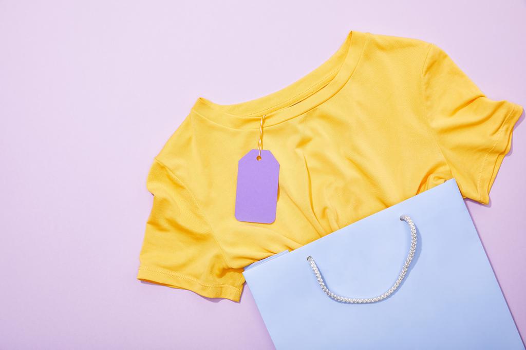 紫色の買い物袋に紫色の紙の販売タグを持つ黄色のTシャツのトップビュー  - 写真・画像