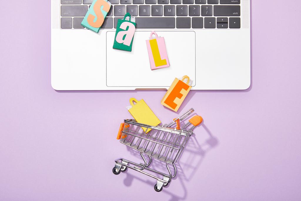 Draufsicht auf bunte Spielzeugtaschen mit Verkauf Schriftzug auf Laptop fällt in Spielzeugwarenkorb auf violett  - Foto, Bild