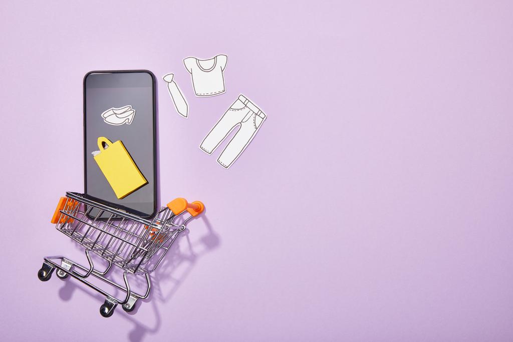 top view of smartphone with paper cut of clothes and yellow paper bag falling into toy cart on violet  - Photo, Image