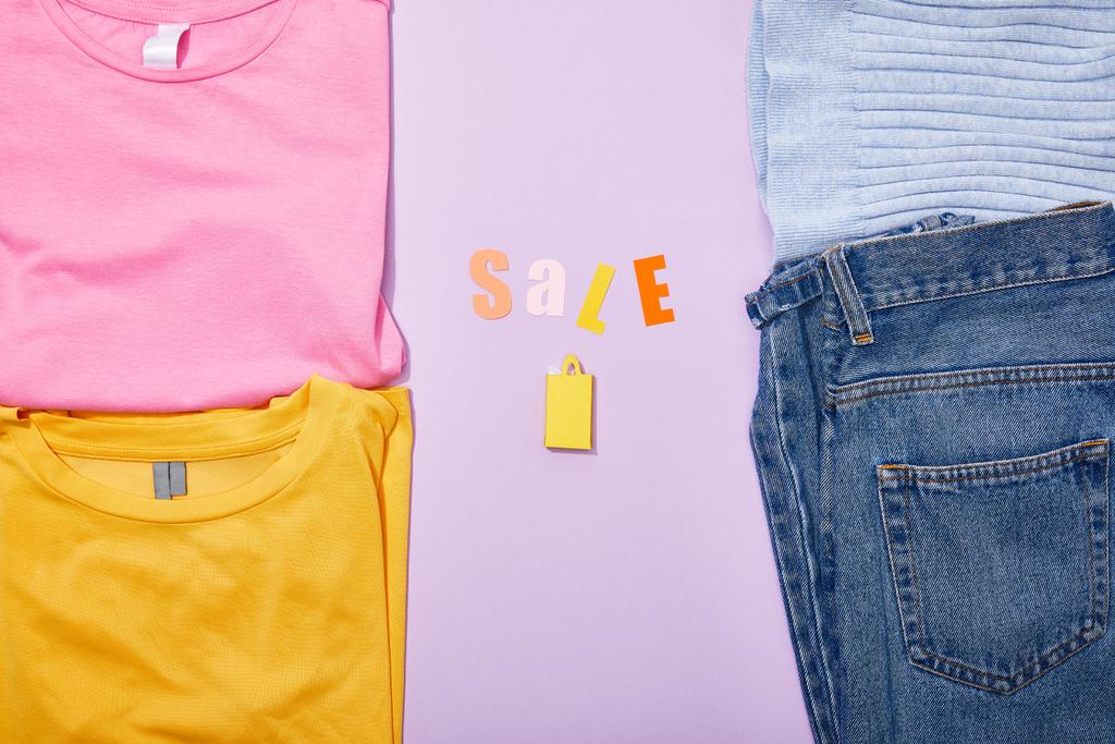 bovenaanzicht van de verkoop belettering boven winkelen papieren zak in de buurt van kleurrijke kleren op Violet  - Foto, afbeelding