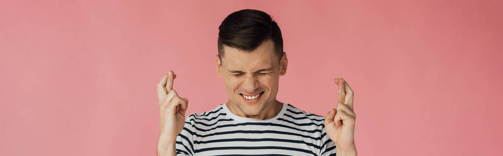panoramic shot of front view of smiling man in striped t-shirt with closed eyes and crossed fingers isolated on pink - Photo, Image