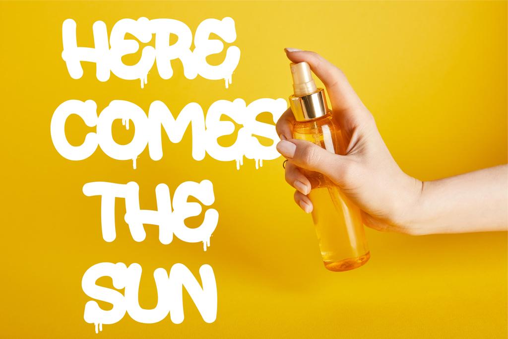 περικομμένη όψη της γυναίκας που κρατά το μπουκάλι με αντηλιακό σπρέι σε κίτρινο φόντο με εδώ έρχεται η γραφή του ήλιου - Φωτογραφία, εικόνα