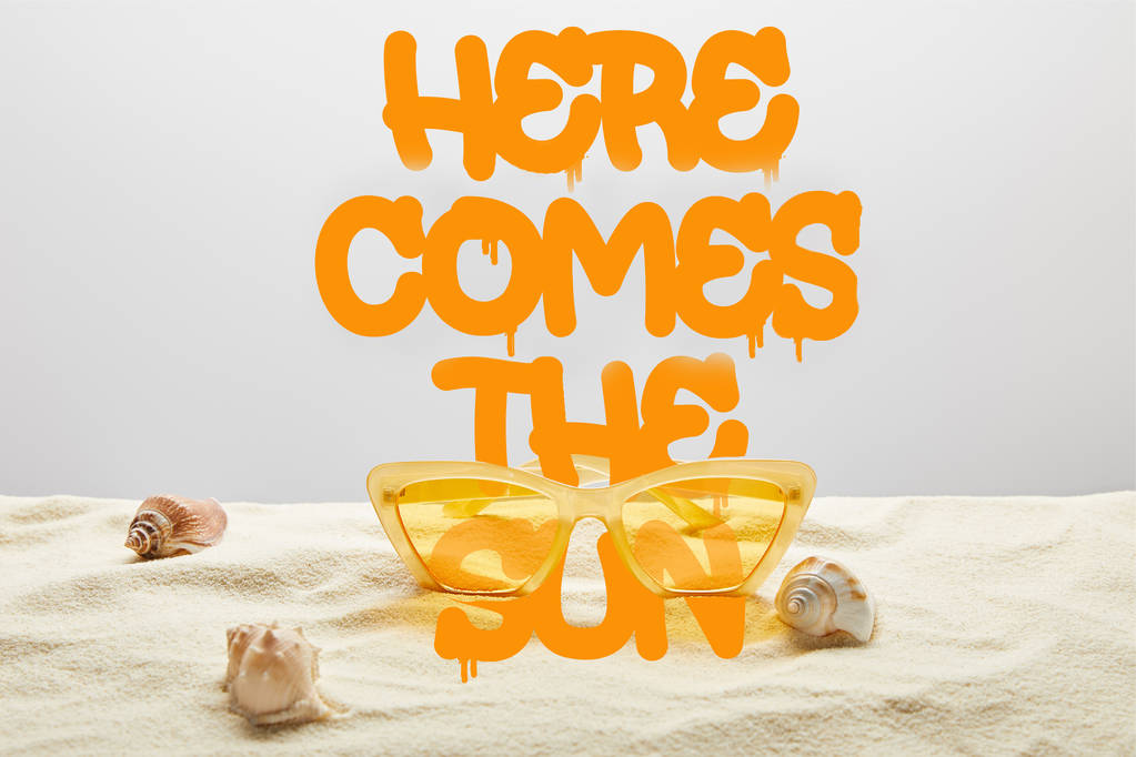 yellow stylish sunglasses on sand with seashell on grey background with here comes the sun lettering - Photo, Image