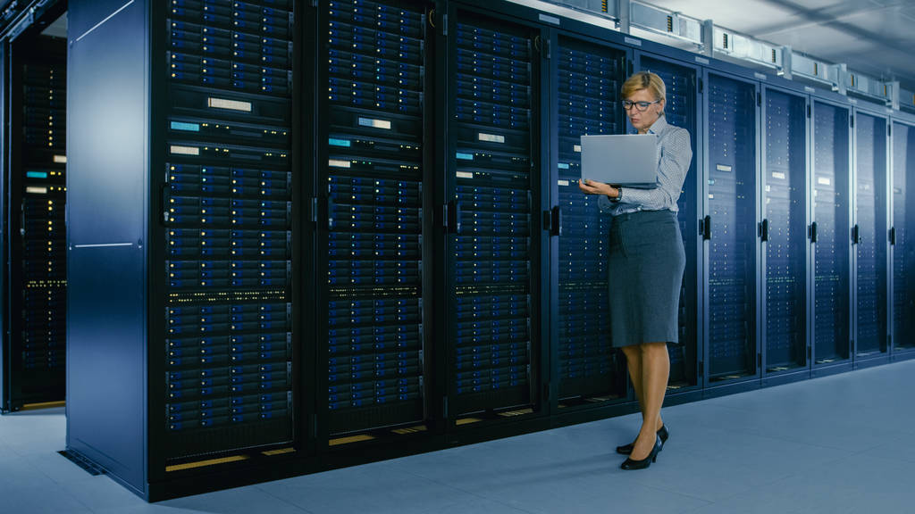 W centrum danych: Kobieta IT technika stoi w pobliżu szafy Rack i uruchamia program konserwacji na laptopie, kontroluje operacyjne optymalne funkcjonowanie serwera. - Zdjęcie, obraz