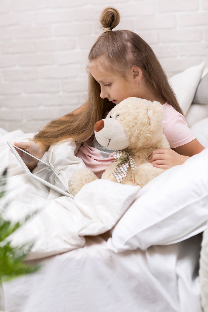 かわいい小さな女の子は、ベッドに横たわってデジタルタブレットを使用しています. - 写真・画像