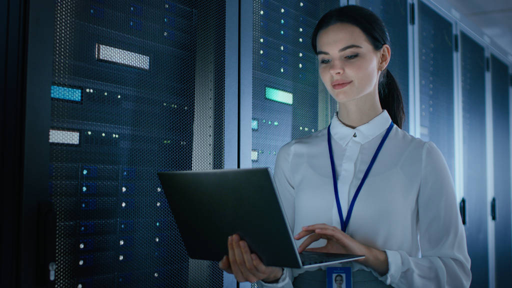 Pewny siebie i piękna kobieta IT Engineer pracuje na laptopie w centrum danych stojąc przed rack Server. Uruchamianie diagnostyki lub wykonywanie prac konserwacyjnych. - Zdjęcie, obraz
