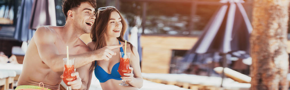 Panoramaaufnahme eines fröhlichen jungen Mannes, der mit dem Finger zeigt, während er neben seiner hübschen Freundin mit einem Glas Erfrischungsgetränk sitzt - Foto, Bild