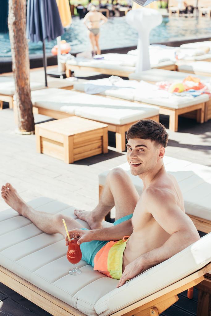 handsome young man smiling at camera while resting on deck chair with glass of refreshing beverage - Photo, Image
