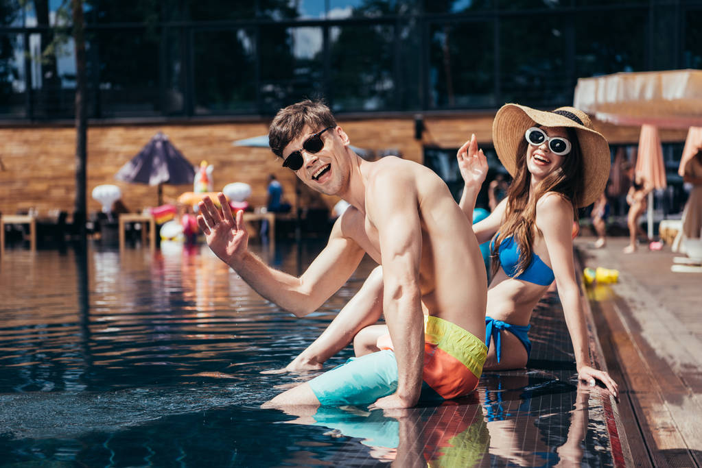 Wesoła Młoda para w okularach przeciwsłonecznych machając rękami przy kamerze podczas relaksu przy basenie - Zdjęcie, obraz