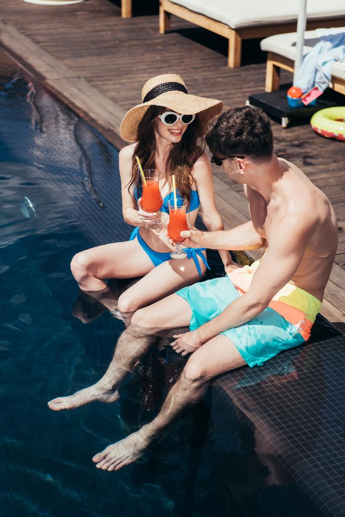 プールサイドに座っている間、幸せな若いカップルは、さわやかな飲み物で眼鏡を鳴らします - 写真・画像