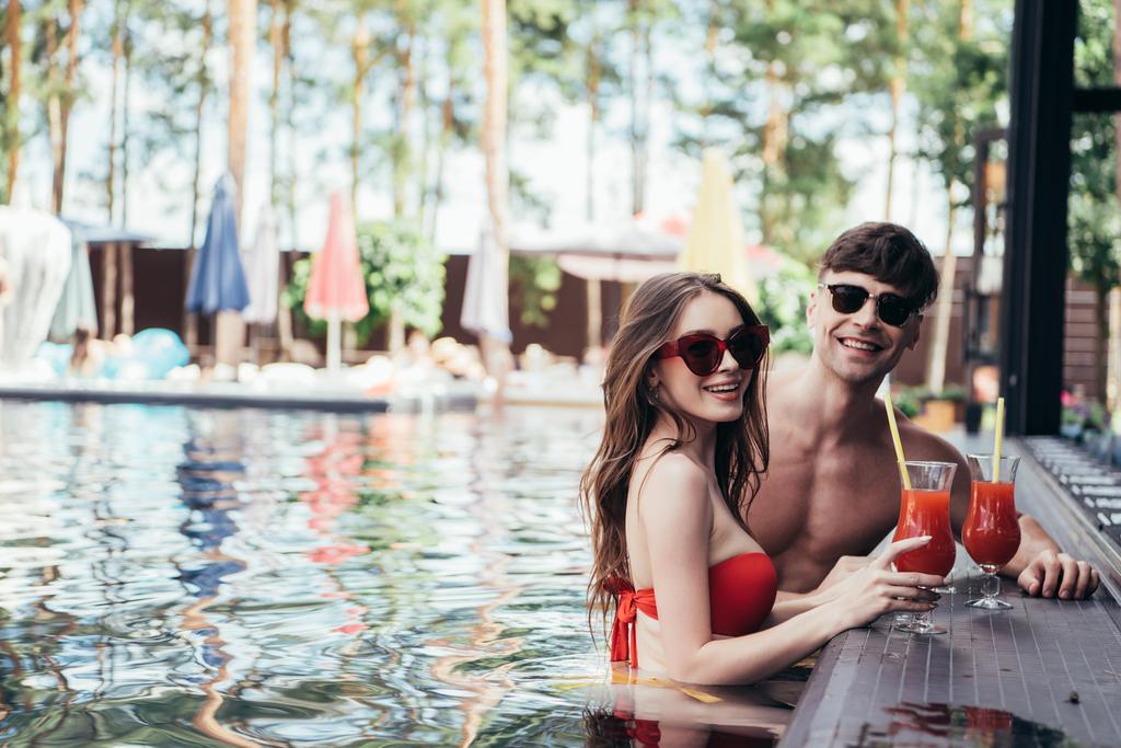 happy young couple relaxing in swimming pool with glases of refreshing drink an smiling at camera - Photo, Image
