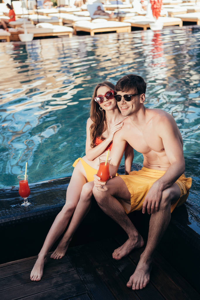 happy young woman hugging handsome boyfriend while sitting at poolside together - Photo, Image