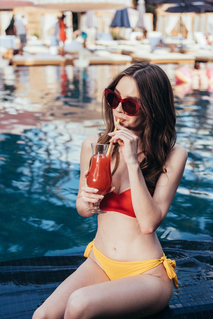 krásná mladá žena v plavkách a slunečních brýlích popíjí osvěžující nápoj při sezení na poolové straně - Fotografie, Obrázek