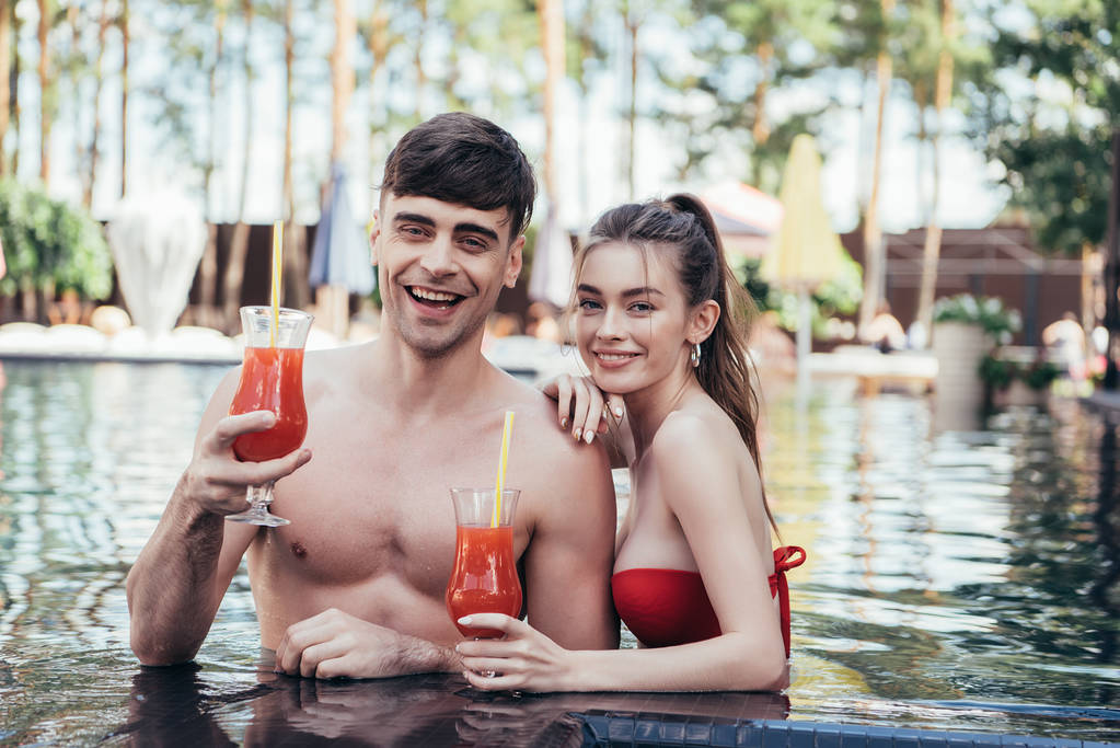 Szczęśliwa Młoda para uśmiechając się do kamery podczas relaksu w basenie z szklankami orzeźwiających napojów - Zdjęcie, obraz