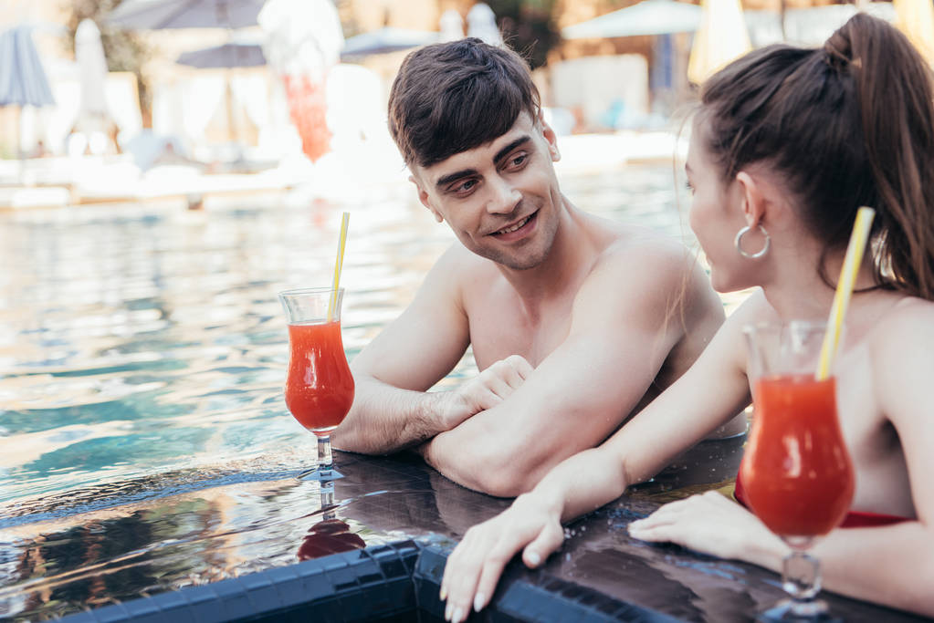 vidám fiatal pár nézi egymást, míg a medence melletti pihenő pohár frissítő italt - Fotó, kép