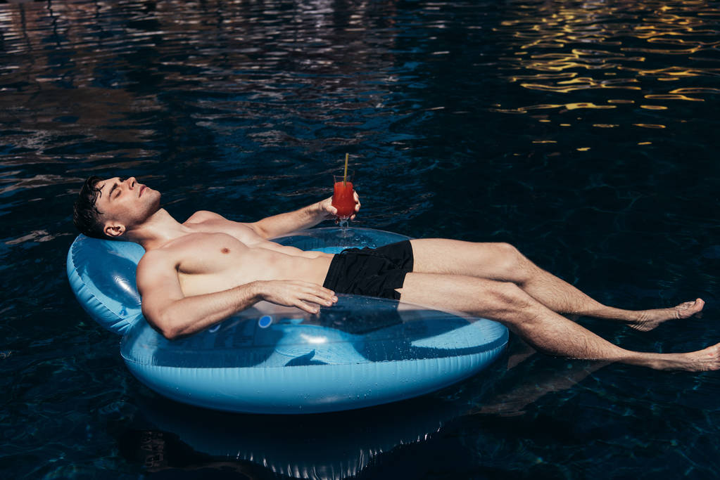 przystojny młody człowiek opalanie się na pierścień pływacki w basenie z kieliszek orzeźwiającego napoju - Zdjęcie, obraz