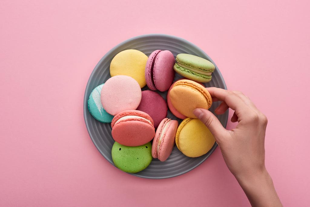 cropped view of female hand near plate with multicolored delicious French macaroons on pink background - Photo, Image