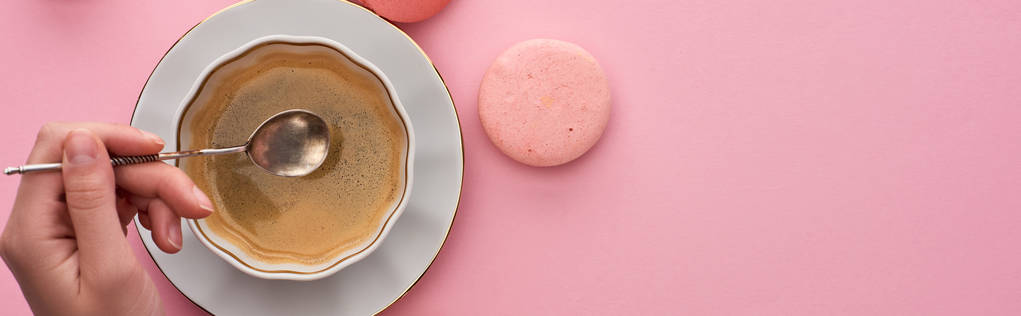 vista cortada da mulher segurando colher de prata acima da xícara de café perto de deliciosos macaroons franceses no fundo rosa, tiro panorâmico
 - Foto, Imagem