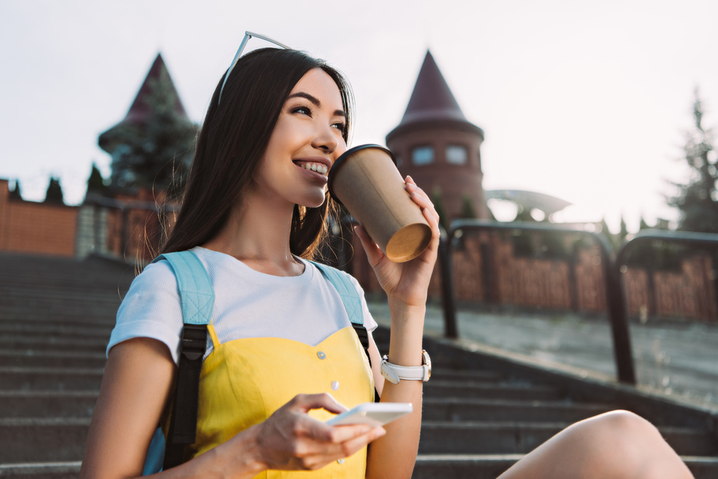 sonriente y asiático mujer sosteniendo smartphone y bebiendo café
  - Foto, imagen