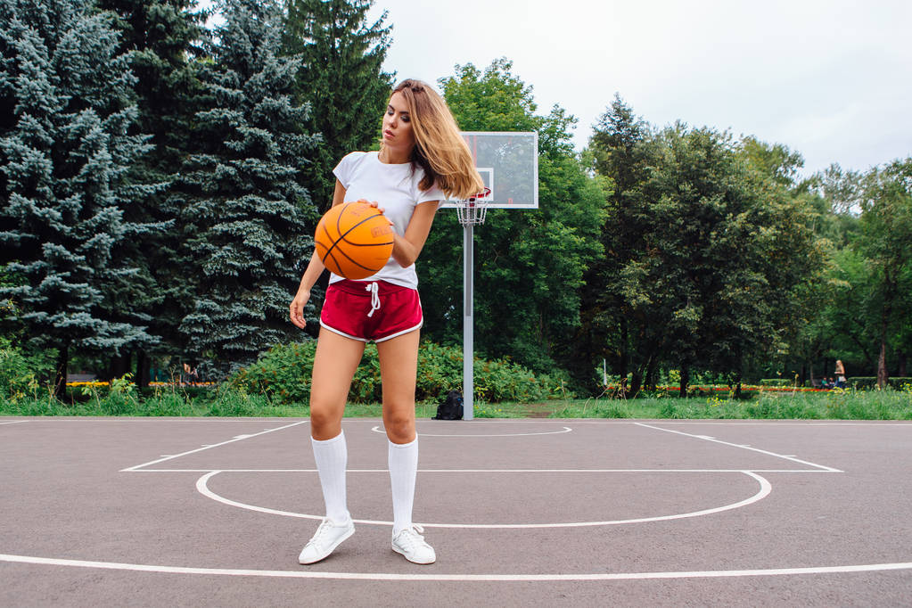 Hermosa joven vestida con camiseta blanca, pantalones cortos y zapatillas de deporte, juega con una pelota en una cancha de baloncesto
. - Foto, imagen