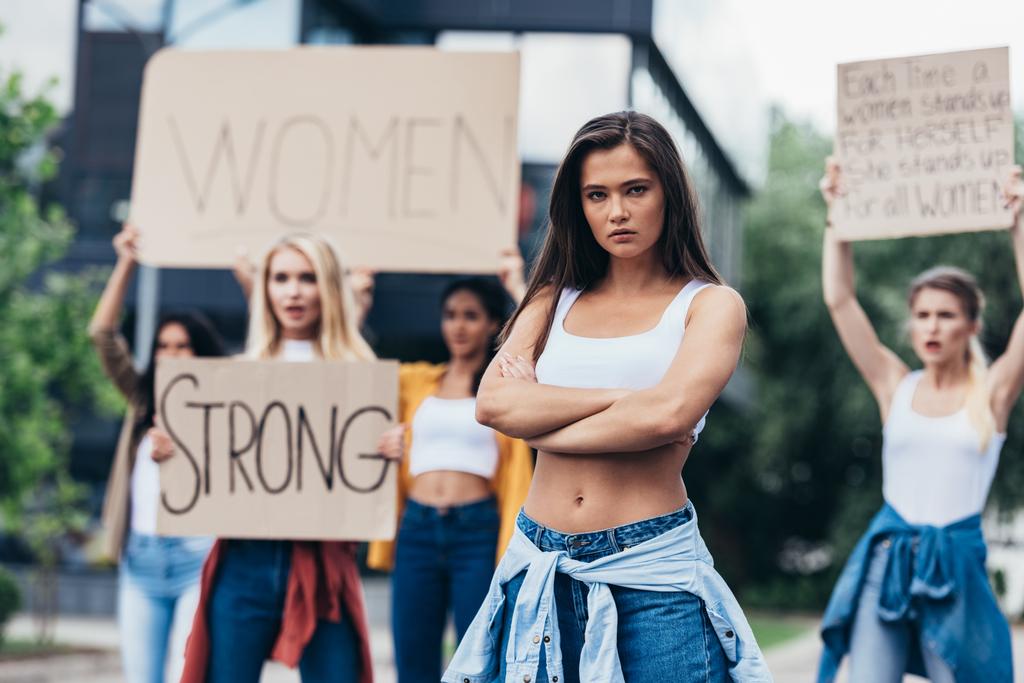 sokakta feminist sloganlar taşıyan pankartlar tutan kadınların yanında kolları kapalı duran ciddi feminist ayakta - Fotoğraf, Görsel