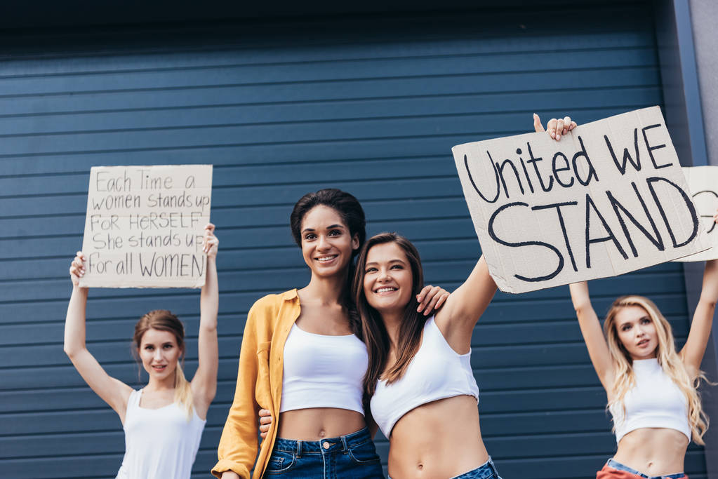 cztery uśmiechnięte wieloetniczne feministki obejmujące i trzymające tablice z sloganami na ulicy - Zdjęcie, obraz