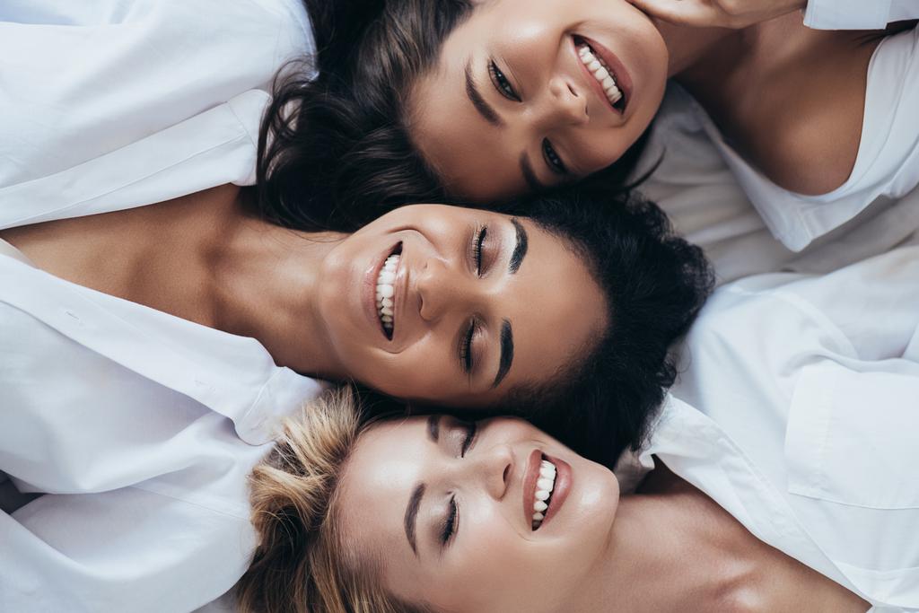 Widok z góry na trzy uśmiechnięte wieloetniczne kobiety w białych koszulkach leżących z zamkniętymi oczami - Zdjęcie, obraz