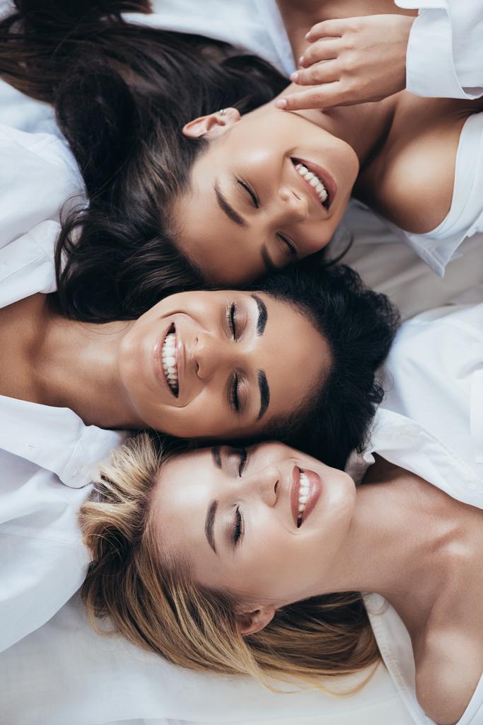 вид сверху на трех улыбающихся многонациональных женщин в белых рубашках с закрытыми глазами
 - Фото, изображение
