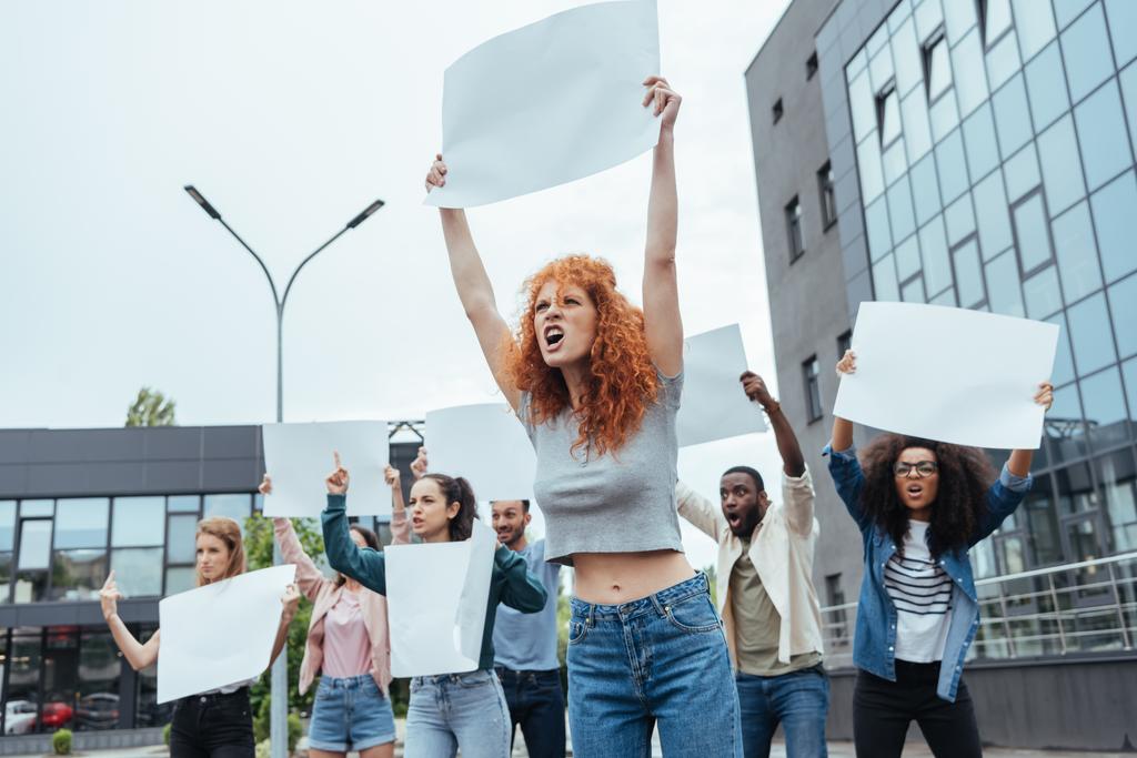 selectieve focus van Angry Redhead meisje houden blanco plakkaat op vergadering in de buurt van multiculturele mensen  - Foto, afbeelding