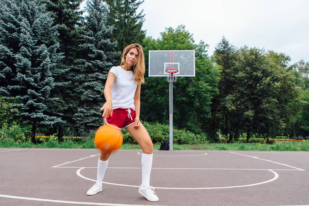 schönes junges Mädchen in weißem T-Shirt, kurzer Hose und Turnschuhen, spielt mit einem Ball auf einem Basketballfeld. - Foto, Bild