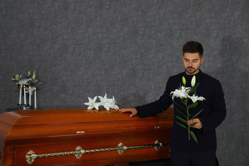 Сумний молодий чоловік з білими ліліями біля коробки в похоронному будинку
 - Фото, зображення