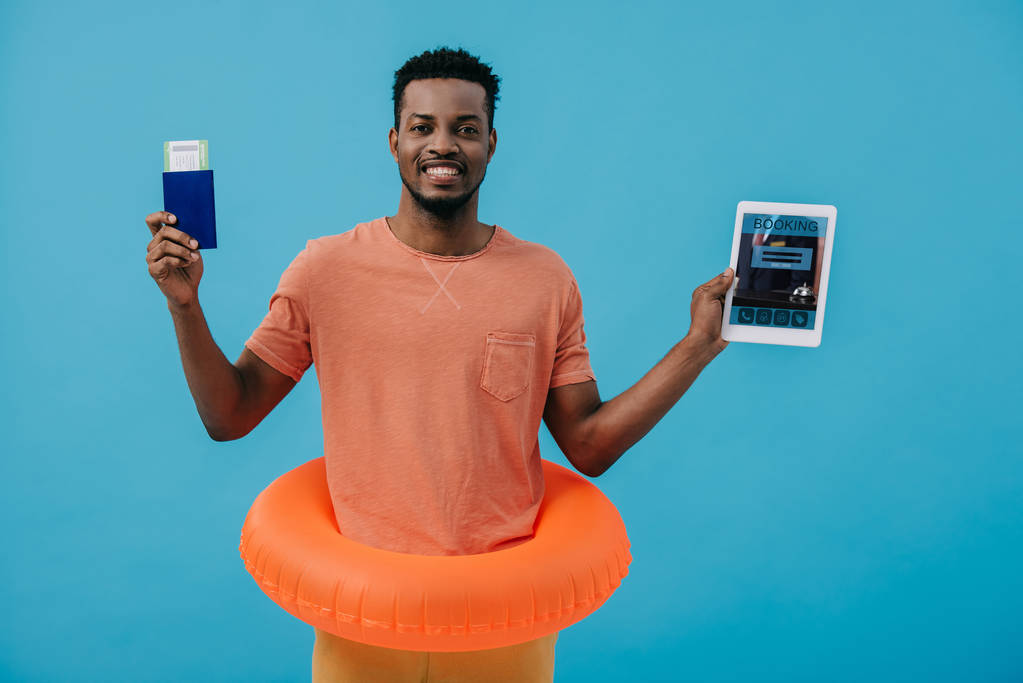KYIV, UKRAINE - JUNHO 27, 2019: homem afro-americano alegre com passaporte e tablet digital com aplicativo de reserva na tela isolado em azul
  - Foto, Imagem