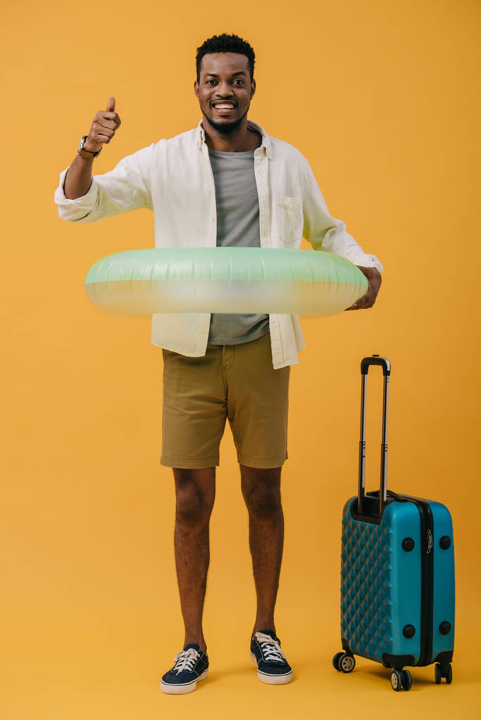 vrolijke Afro-Amerikaanse man staand met zwem ring en bagage terwijl duim omhoog wordt weergegeven op oranje - Foto, afbeelding