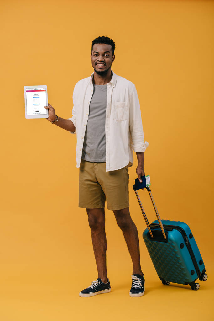 KYIV, UCRANIA - 27 de junio de 2019: alegre afroamericano de pie con el equipaje y la celebración de la tableta digital con aplicación instagram en la pantalla en naranja
 - Foto, imagen