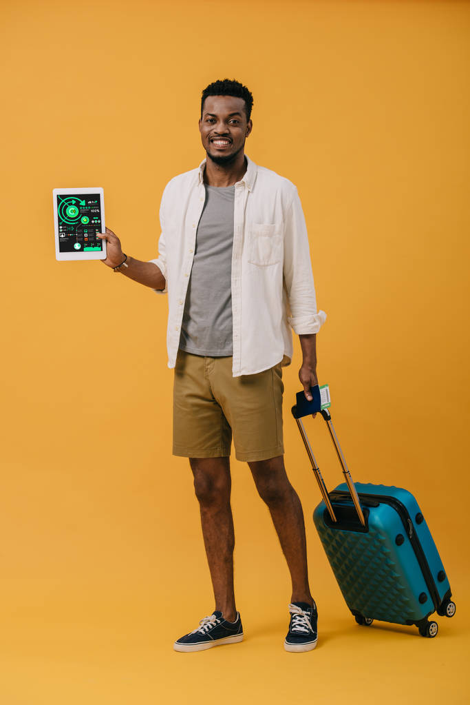 χαρούμενος Αφρικανός Αμερικανός άνθρωπος στέκεται με τις αποσκευές και κρατώντας ψηφιακό δισκίο με διαγράμματα και γραφήματα στην οθόνη για πορτοκαλί - Φωτογραφία, εικόνα