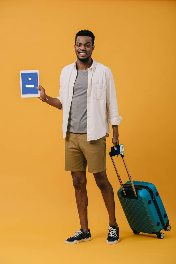 KYIV, UKRAINE kesäkuu 27, 2019: iloinen afrikkalainen amerikkalainen mies seisoo matkatavaroiden kanssa ja pitää digitaalista tablettia facebook-sovelluksella näytöllä oranssilla
 - Valokuva, kuva