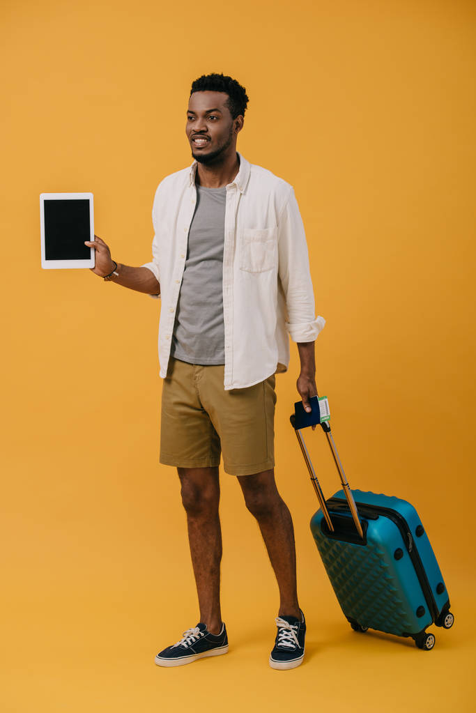 mutlu afrikalı amerikalı adam bagaj ile ayakta ve turuncu boş ekran ile dijital tablet tutan - Fotoğraf, Görsel