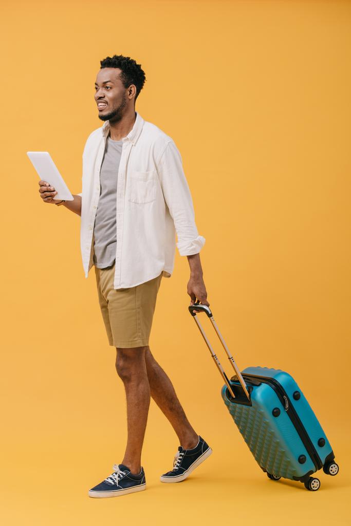 デジタルタブレットを持ち、オレンジ色の荷物を持って歩く幸せなアフリカ系アメリカ人の男性 - 写真・画像