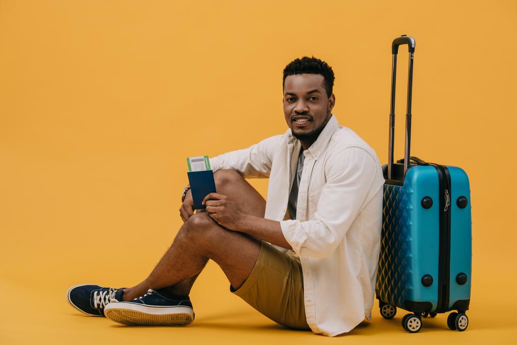 wesoły African American mężczyzna trzyma paszport z biletem lotniczym i siedzi w pobliżu bagażu na pomarańczowo - Zdjęcie, obraz
