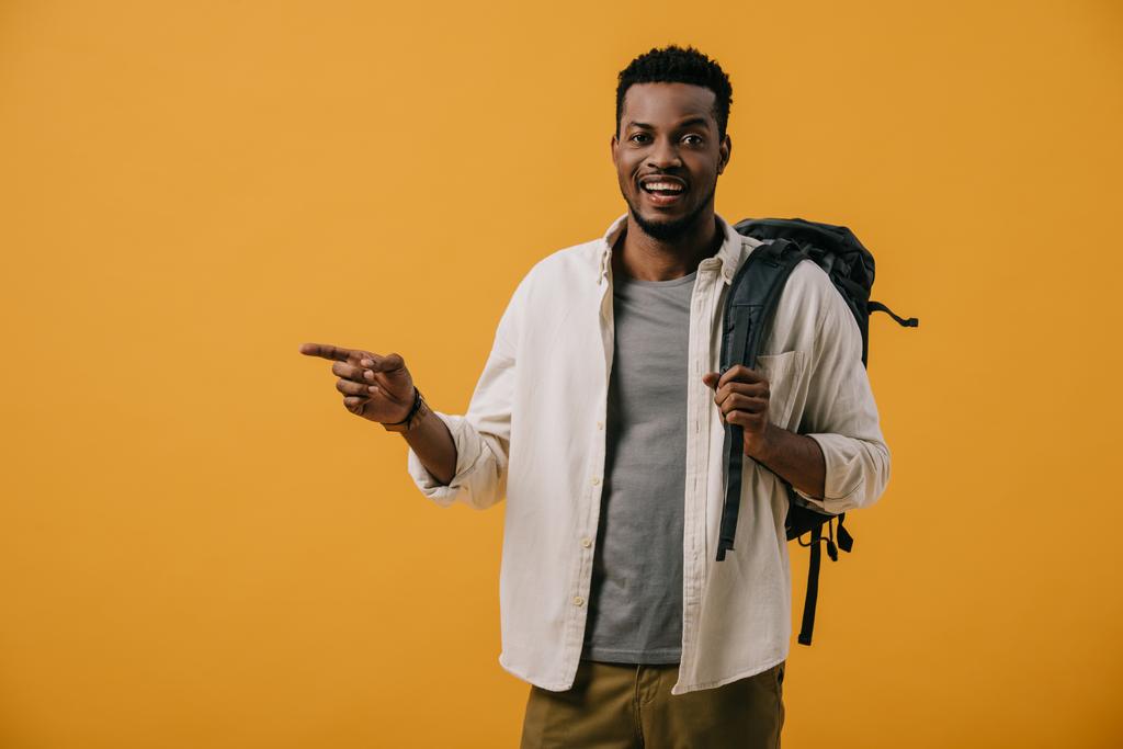 happy african american man pointing with finger while standing with backpack isolated on orange  - Photo, Image