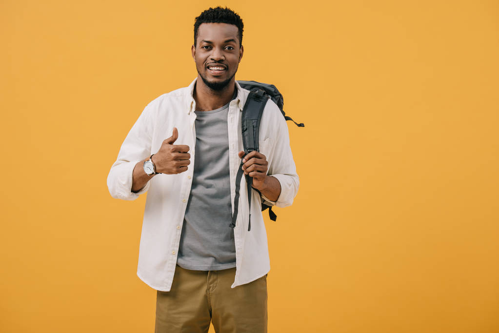 親指を上にしてオレンジ色に孤立したバックパックに触れる幸せなアフリカ系アメリカ人男性  - 写真・画像