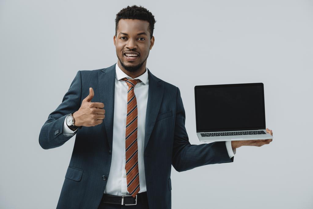 χαρούμενος Αφρικανός Αμερικανός άνθρωπος κρατώντας φορητό υπολογιστή με κενή οθόνη και δείχνοντας αντίχειρας επάνω απομονωμένη σε γκρι  - Φωτογραφία, εικόνα