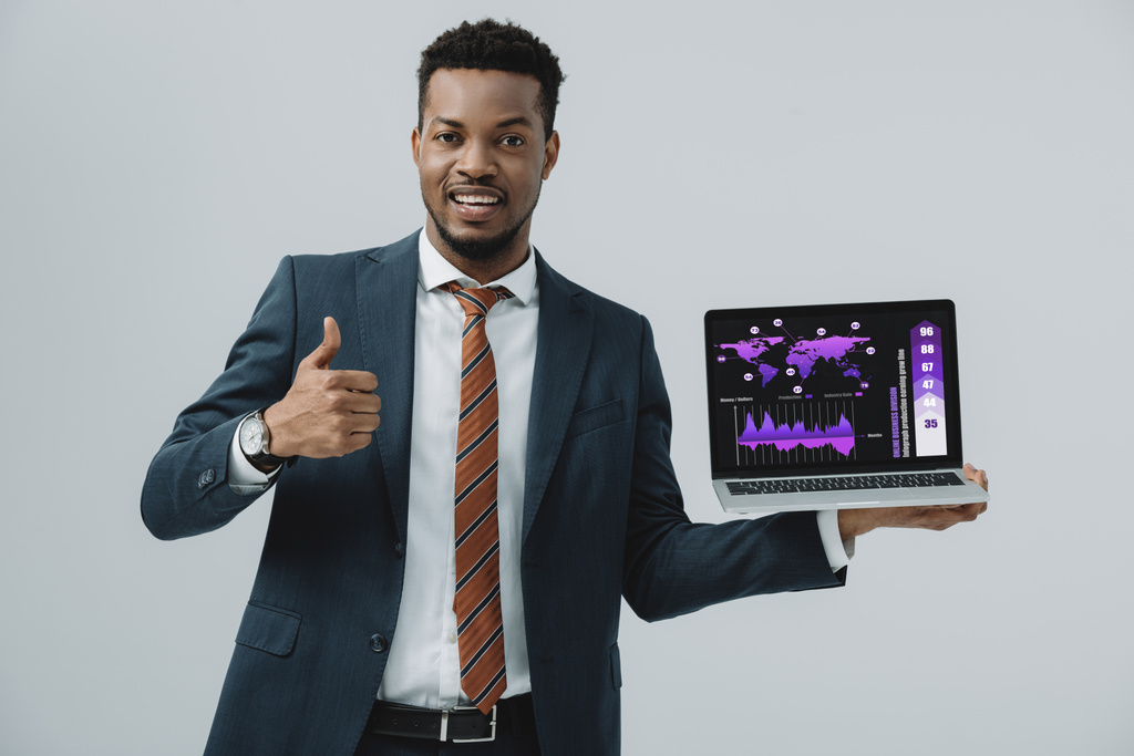vrolijke Afro-Amerikaanse man met laptop met grafieken en grafieken op het scherm en het tonen van duim omhoog geïsoleerd op grijs  - Foto, afbeelding