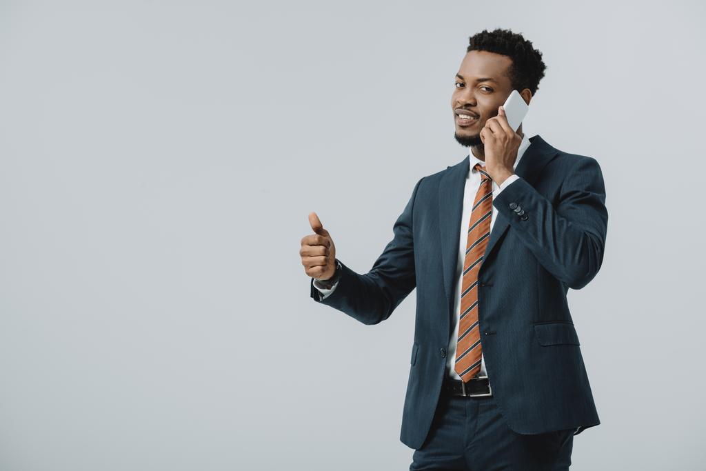 幸せなアフリカ系アメリカ人のビジネスマンは親指を上げて、灰色で隔離されたスマートフォンで話す  - 写真・画像