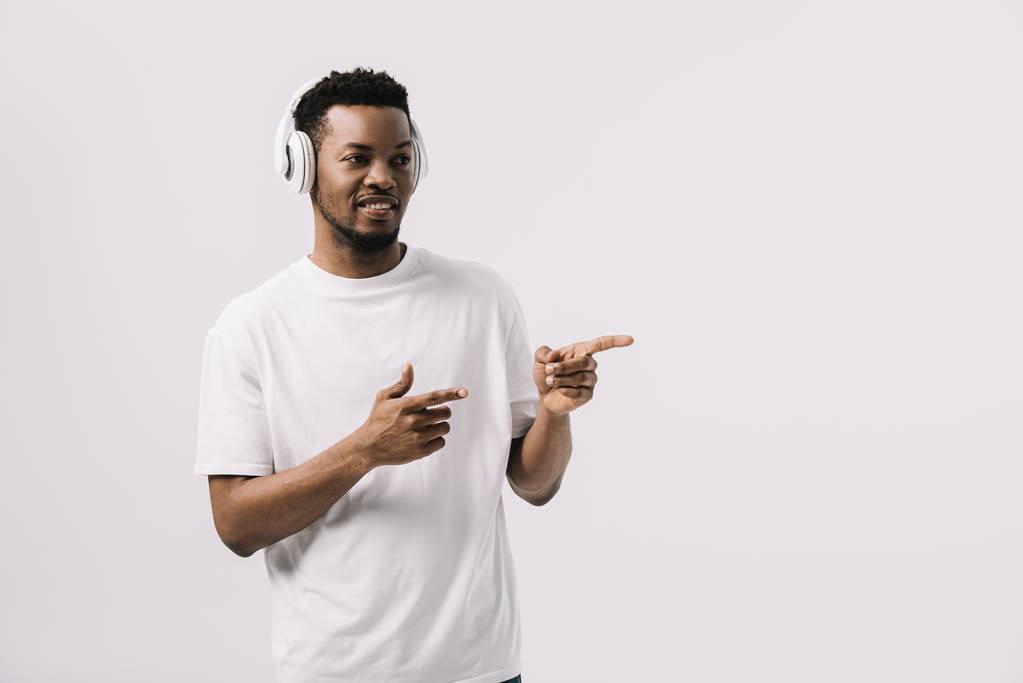 ευτυχισμένος Αφρικανός Αμερικανός άνθρωπος ακούγοντας μουσική στα ακουστικά και δείχνοντας με τα δάχτυλα απομονωμένα σε λευκό  - Φωτογραφία, εικόνα