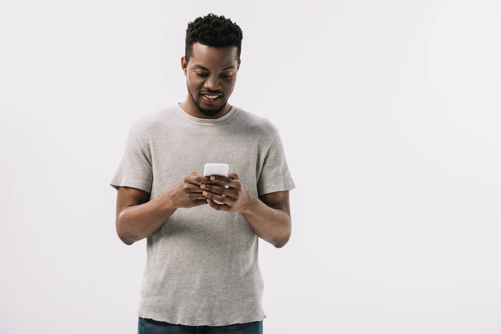 白で隔離されたスマートフォンを使用して灰色のTシャツを着た陽気なアフリカ系アメリカ人男性  - 写真・画像