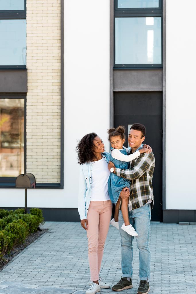 volledige lengte uitzicht van Afro-Amerikaanse familie permanent in de buurt van nieuw huis terwijl vader houden Kid en moeder kijken naar kind - Foto, afbeelding