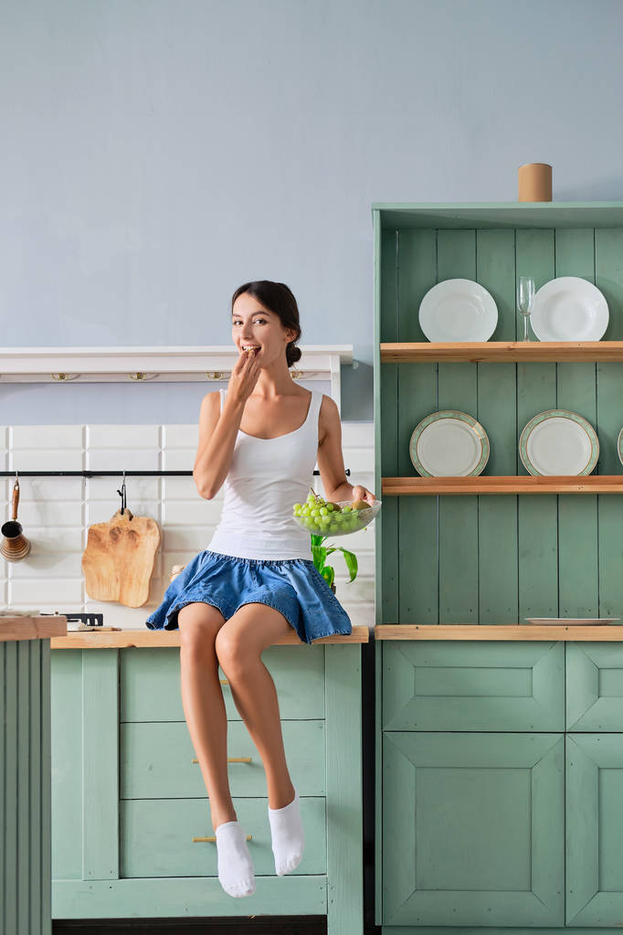 Мила молода жінка сидить на кухонному столі і їсть виноград
 - Фото, зображення